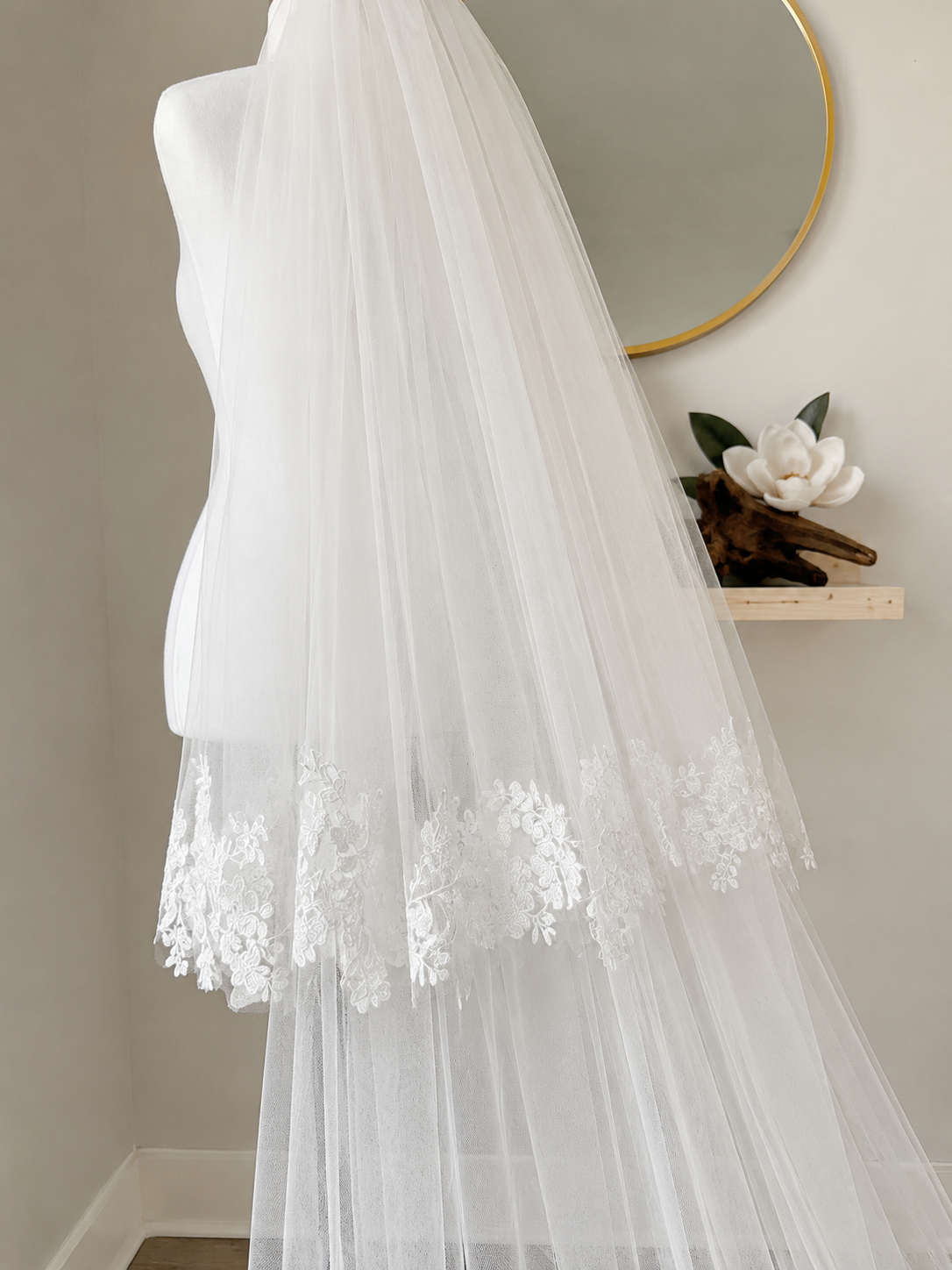 A long white tulle bridal veil with floral lace trim draped over a mannequin torso in an indoor setting with a mirror and shelf in the background.