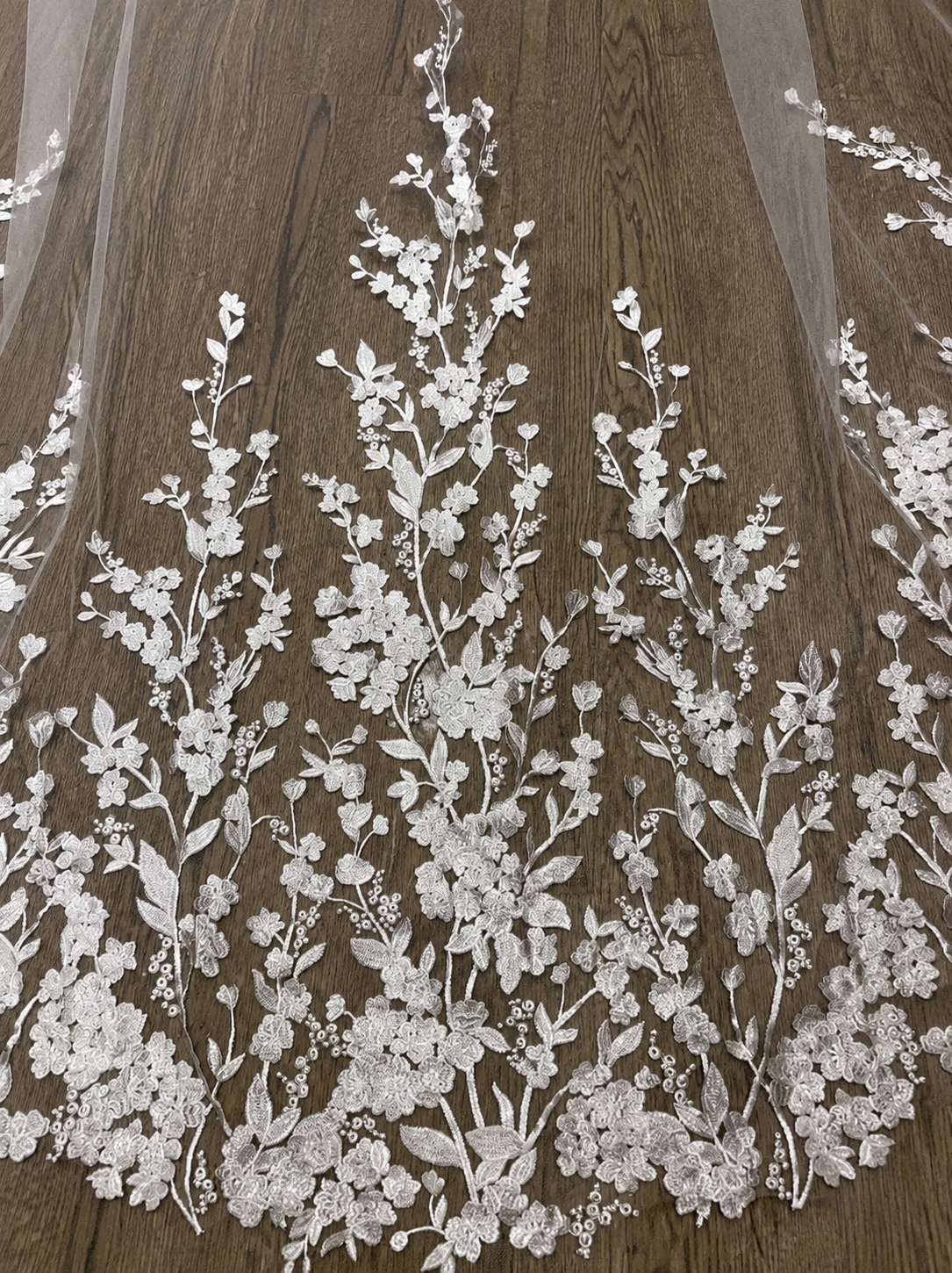 White floral lace applique embroidered on sheer tulle fabric, displayed against a dark brown wood grain background.