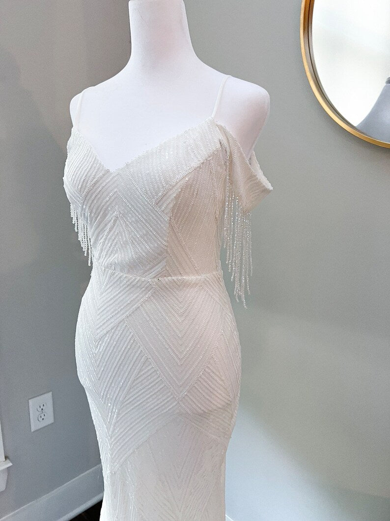 The dress features an off-the-shoulder design with one sleeve draped with beaded fringe, adding a touch of movement and sophistication. The gown is form-fitting with a geometric pattern of sequins and beads that cascade down the entire length, creating a glamorous shimmer effect.