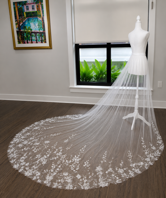 A long white bridal veil with floral lace embroidery spread out on a dark wood floor, displayed on a white dress form mannequin in a room with a window and framed artwork.