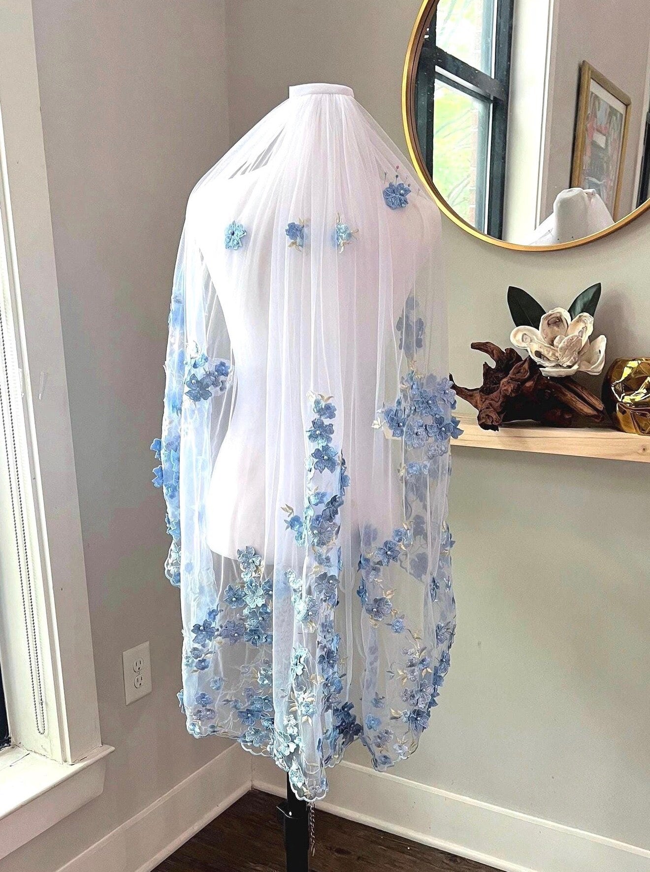 A white bridal veil with light blue floral embroidery draped over a white mannequin torso in an indoor room setting.