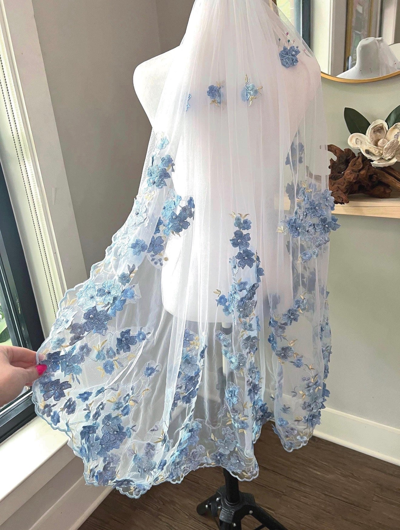 A white tulle bridal veil with blue floral embroidery and applique, displayed on a dress form mannequin indoors. A hand is holding the veil out to the side to show its full length and texture.