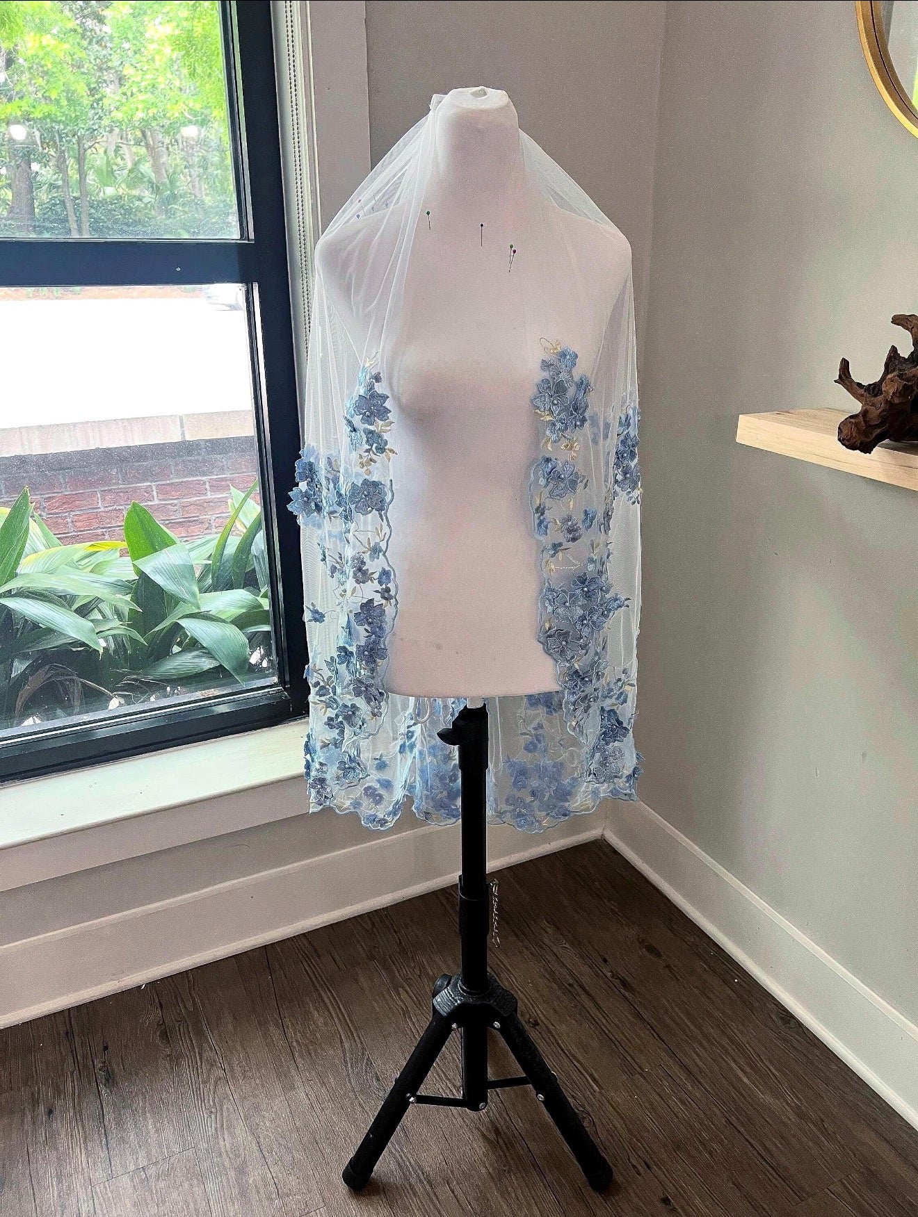 A white bridal veil with light blue floral lace appliqués draped over a white mannequin torso on a black stand, set in a room with a window and wood floor.