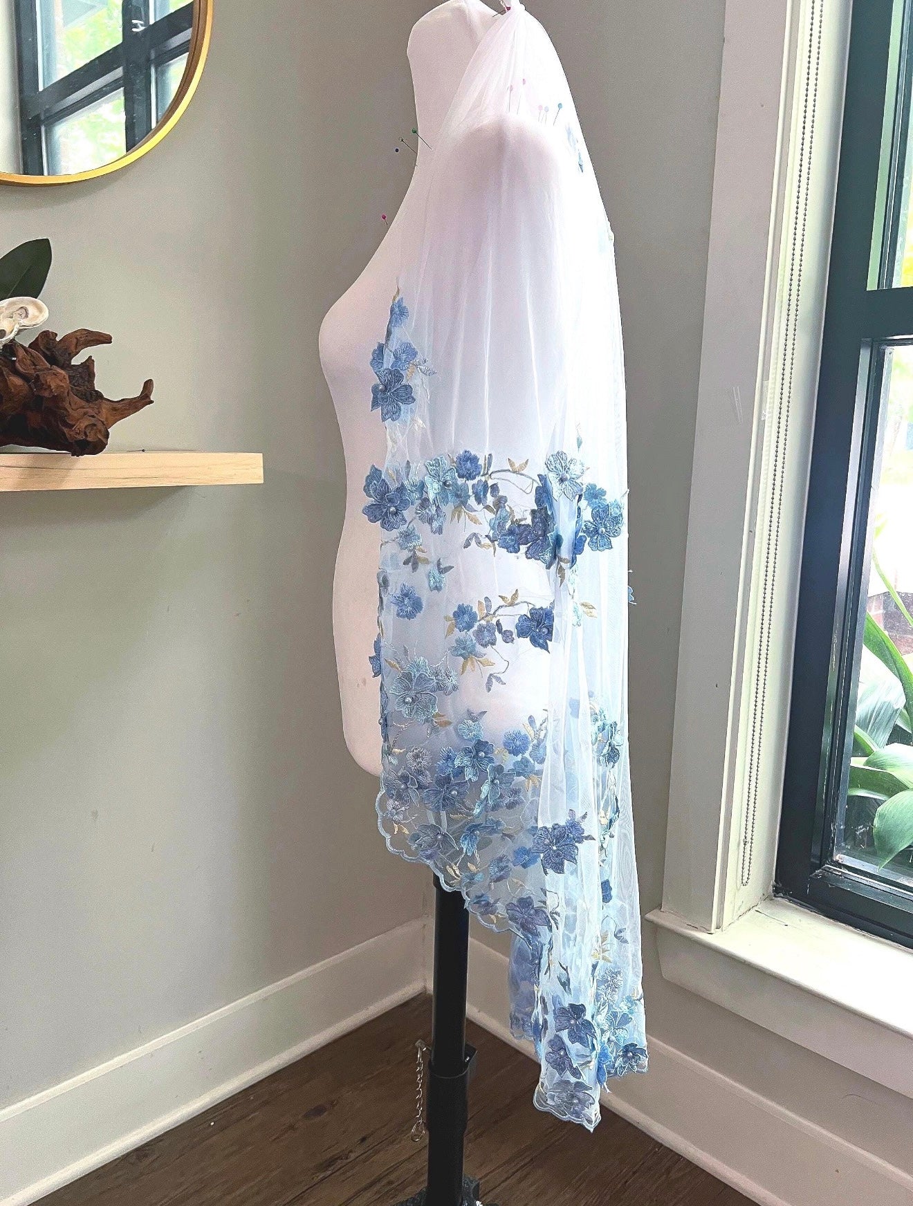 A white tulle bridal veil with blue floral lace applique draped over a white mannequin torso in an indoor room setting.