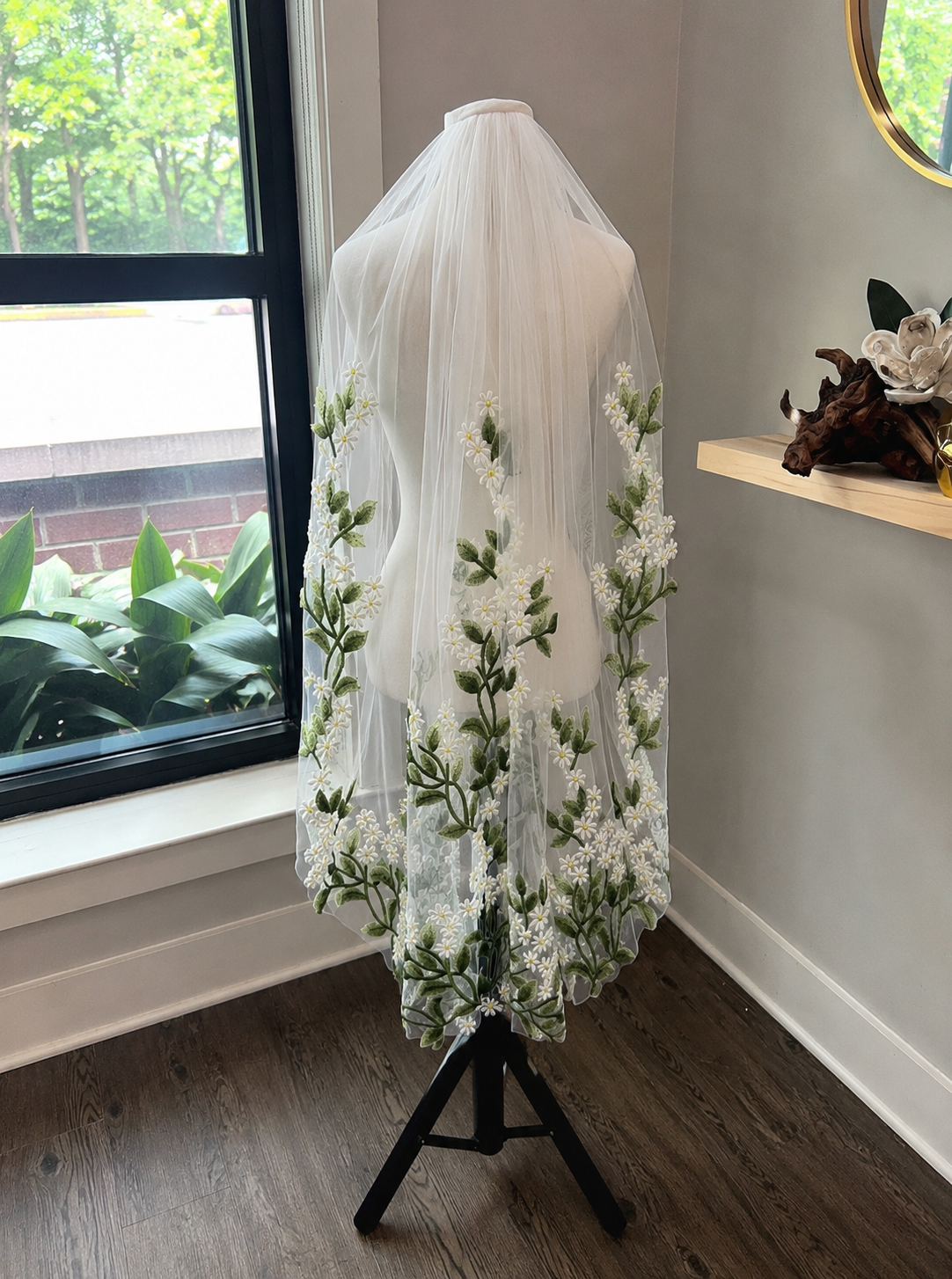 A white bridal veil with vertical floral embroidery of white flowers and green leaves, displayed on a white mannequin torso stand in a room with a window and wood floor.
