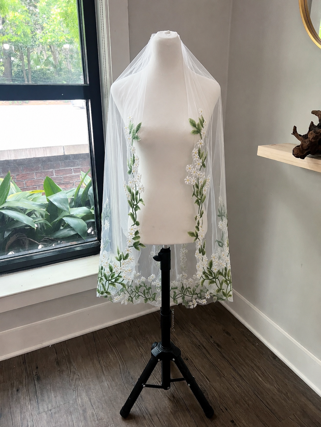 A white bridal veil with green floral embroidery and a floral lace hem, displayed on a white mannequin torso on a black stand in a room with a window and wood floor.