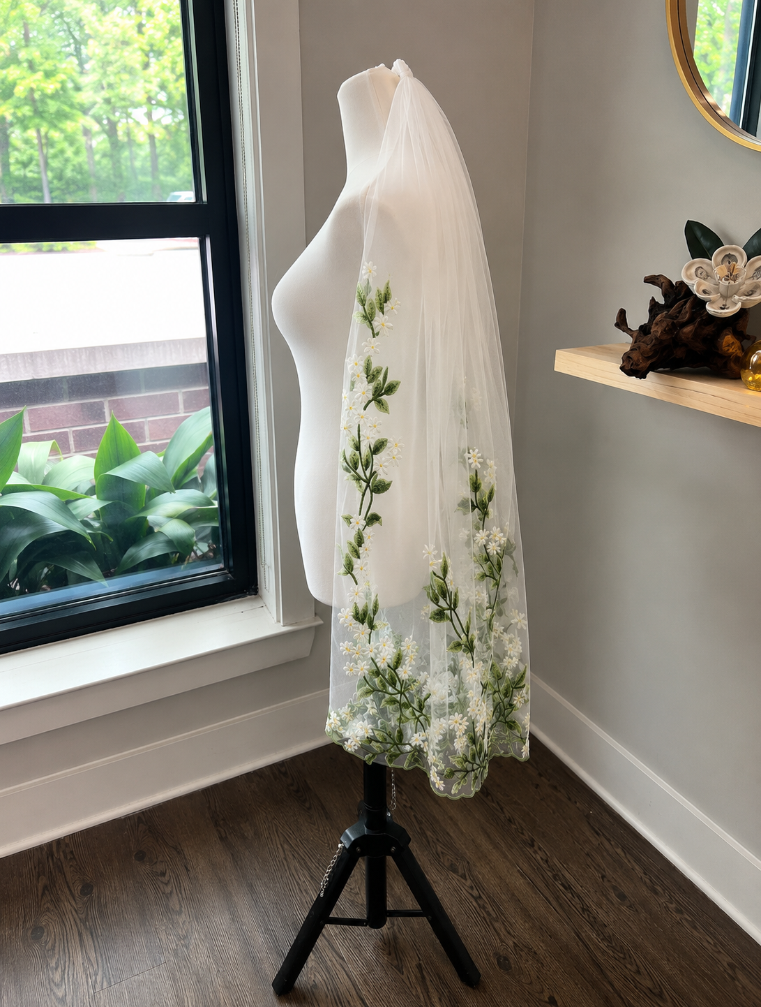 A white tulle bridal veil with green floral embroidery and white lace trim, displayed on a white mannequin torso on a black stand in a room with a window and wood floor.