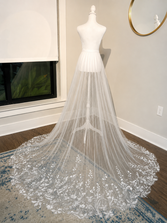 A long white bridal veil with floral lace trim displayed on a white dress form mannequin in an indoor room with wood flooring and a patterned rug.
