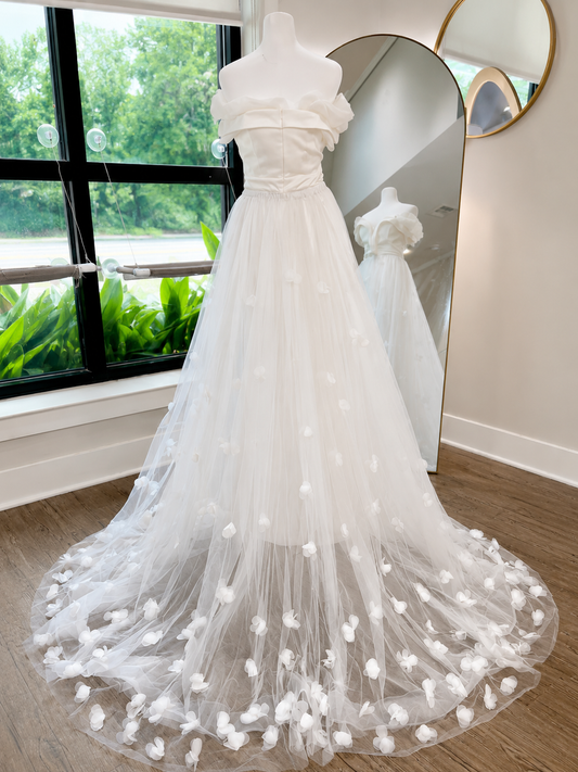 A white off-the-shoulder A-line wedding gown with 3D floral appliqués scattered across the tulle skirt, displayed on a mannequin in a boutique setting with a full-length mirror and window view in the background.