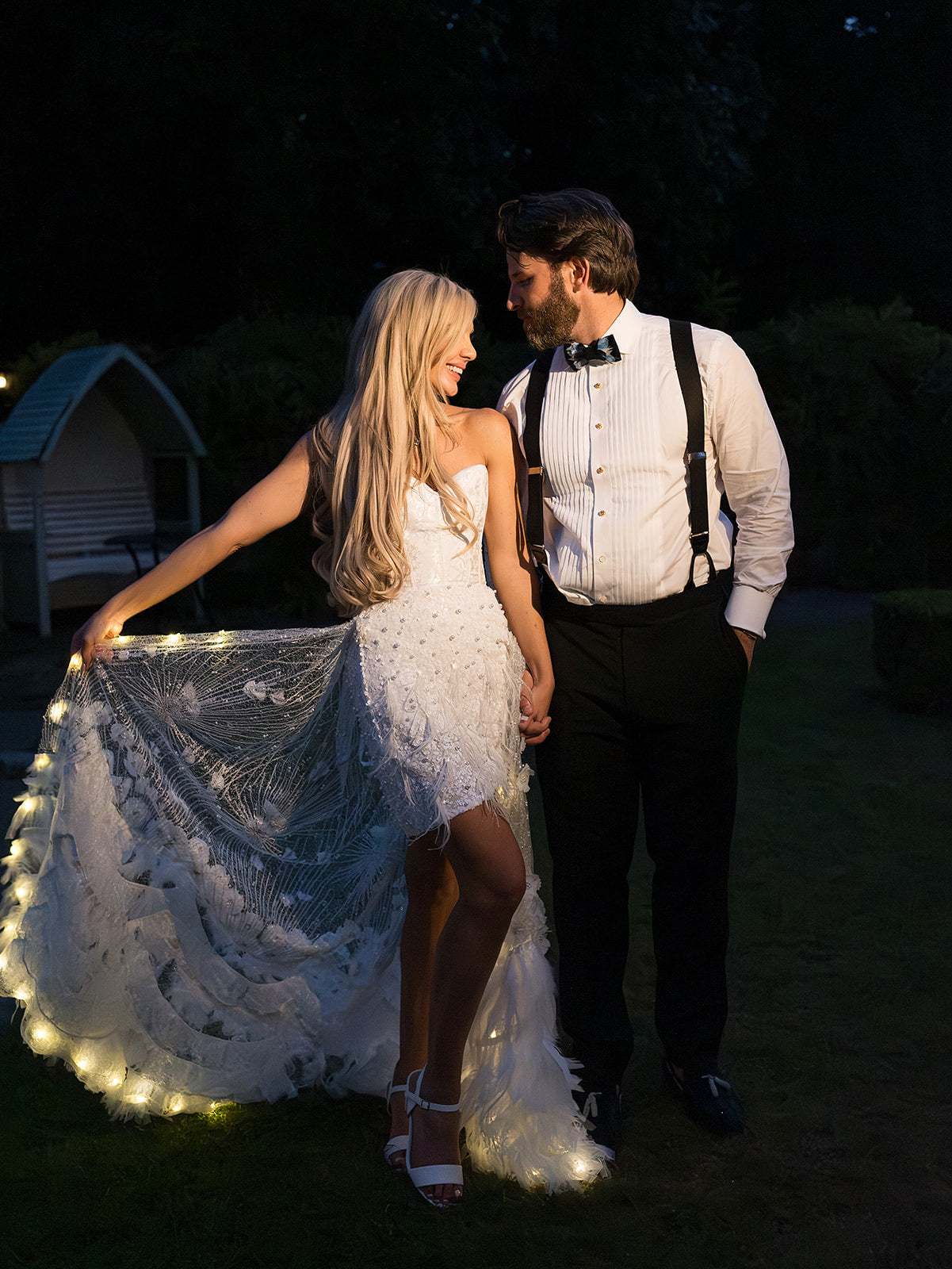 A couple outdoors at night wearing wedding attire. The woman wears a strapless white lace wedding dress with a long ruffled train illuminated by warm string lights along the hem.