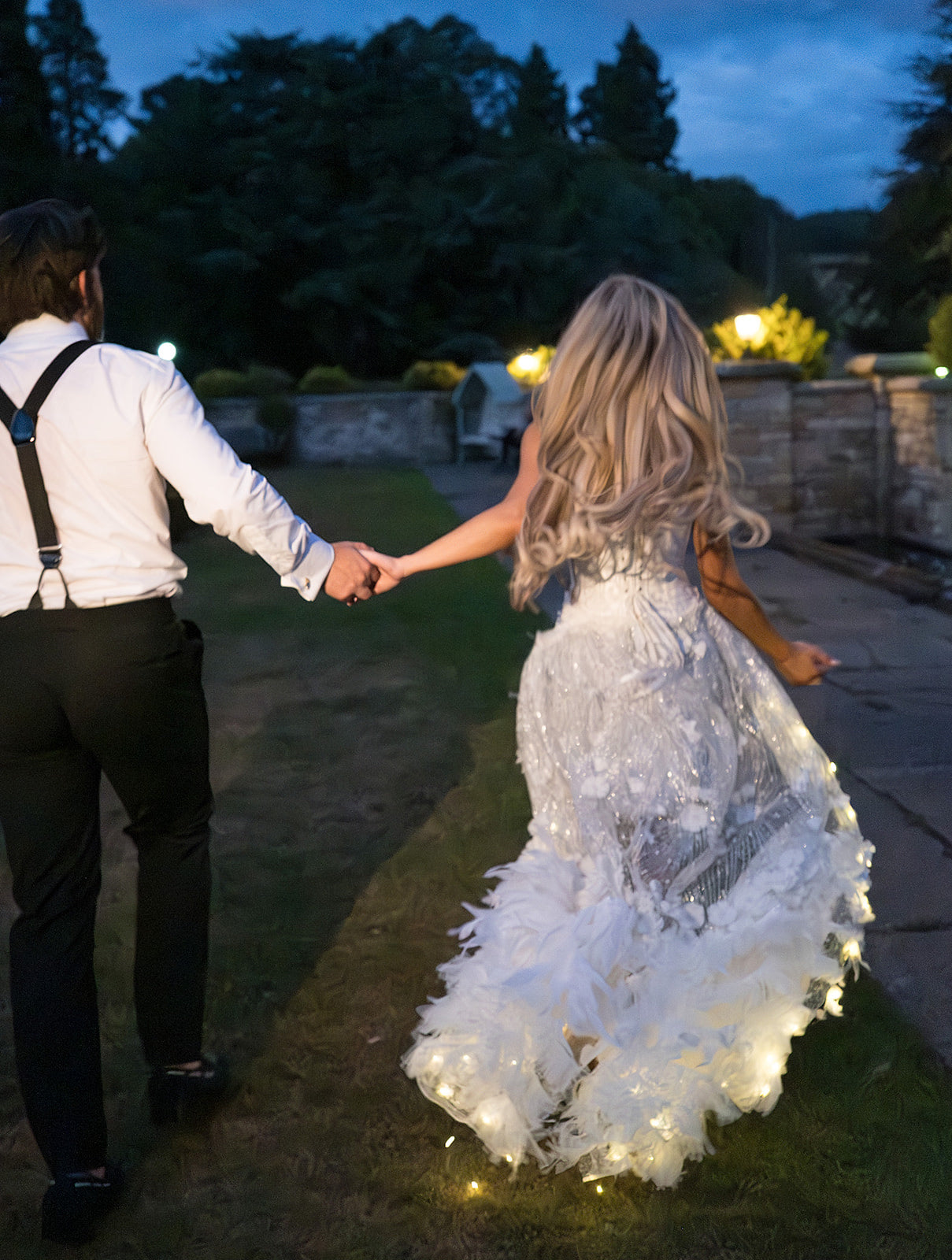 A bride in a white feathered wedding gown with glowing fairy lights woven into the skirt runs and twirls outdoors at dusk while holding hands with a groom in a white shirt and black trousers.