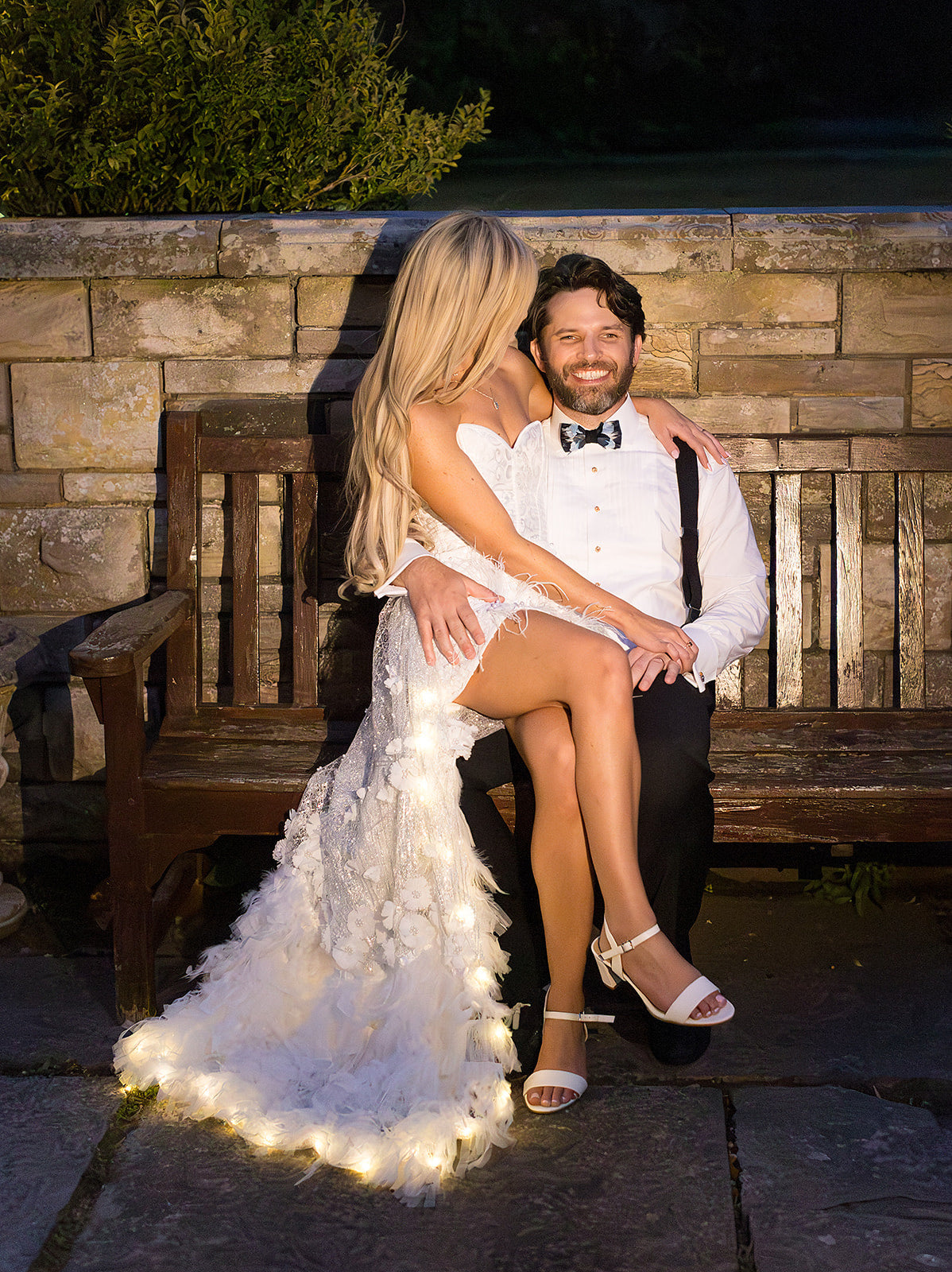 A bride in a white feather-trimmed wedding gown and a groom in a white shirt and black bow tie sit together on a wooden bench outdoors at sunset, with the bride's dress illuminated by warm light.