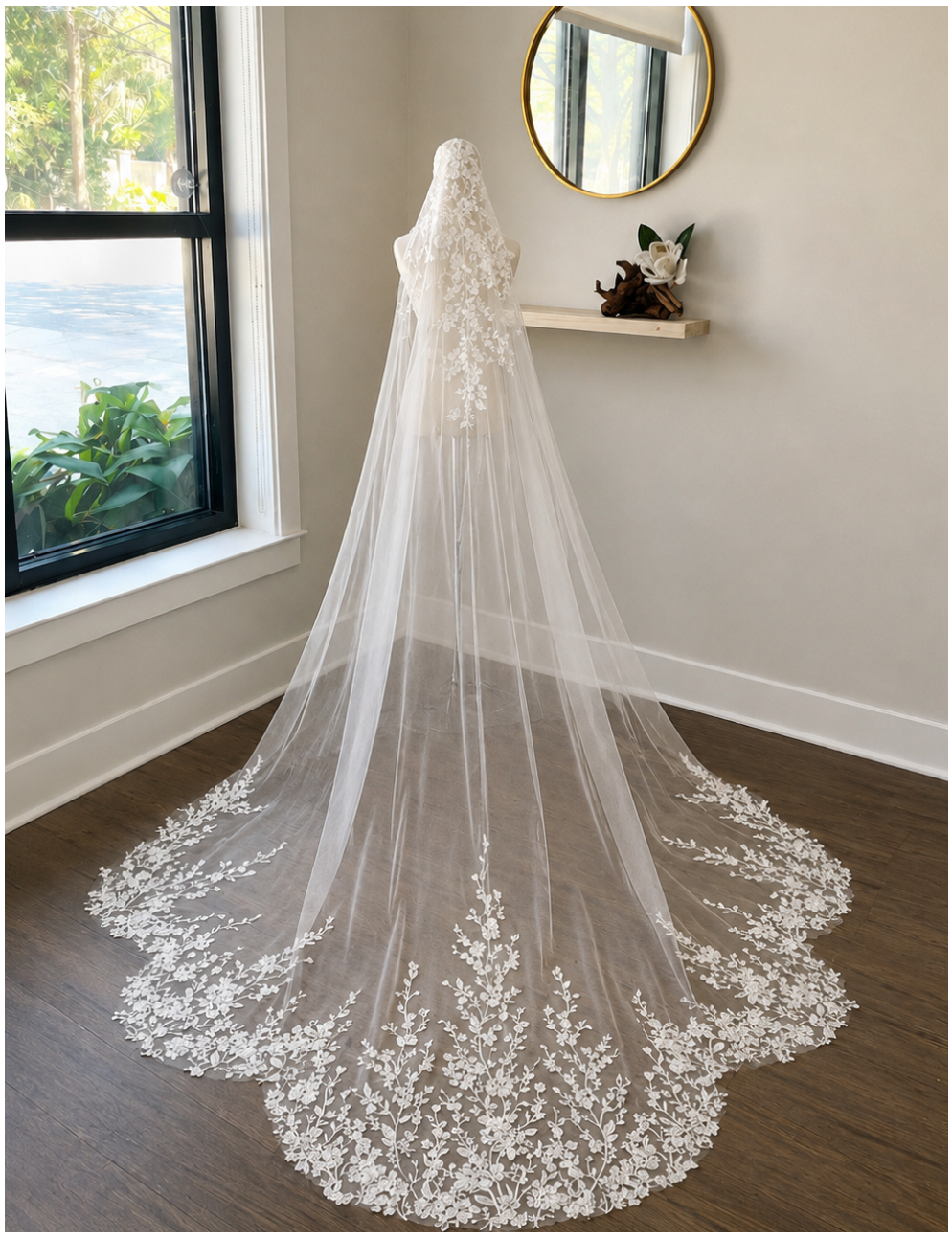 A long white lace bridal veil with floral embroidery and a sheer tulle train, displayed on a dress form in a bright room with a window, wood floor, and wall shelf.