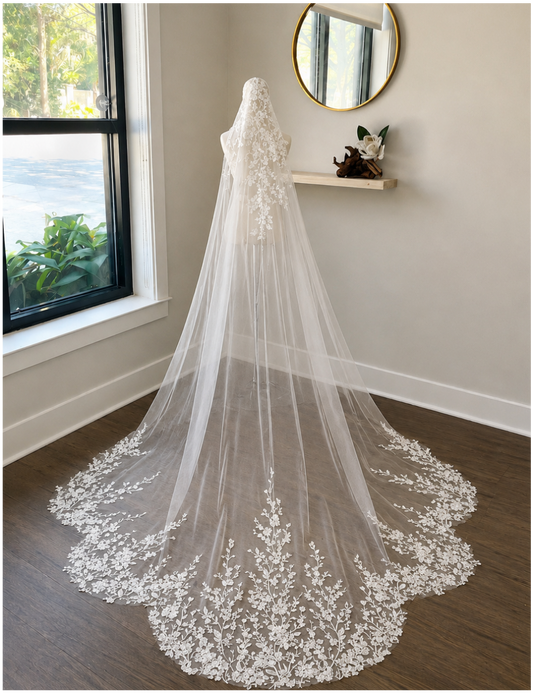A long white lace bridal veil with floral embroidery and a sheer tulle train, displayed on a dress form in a bright room with a window, wood floor, and wall shelf.