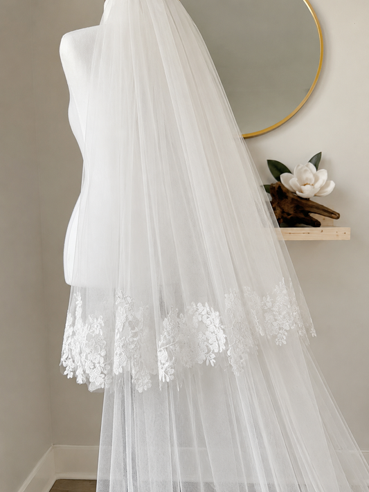A long white tulle bridal veil with floral lace trim draped over a mannequin torso in an indoor setting with a mirror and shelf in the background.
