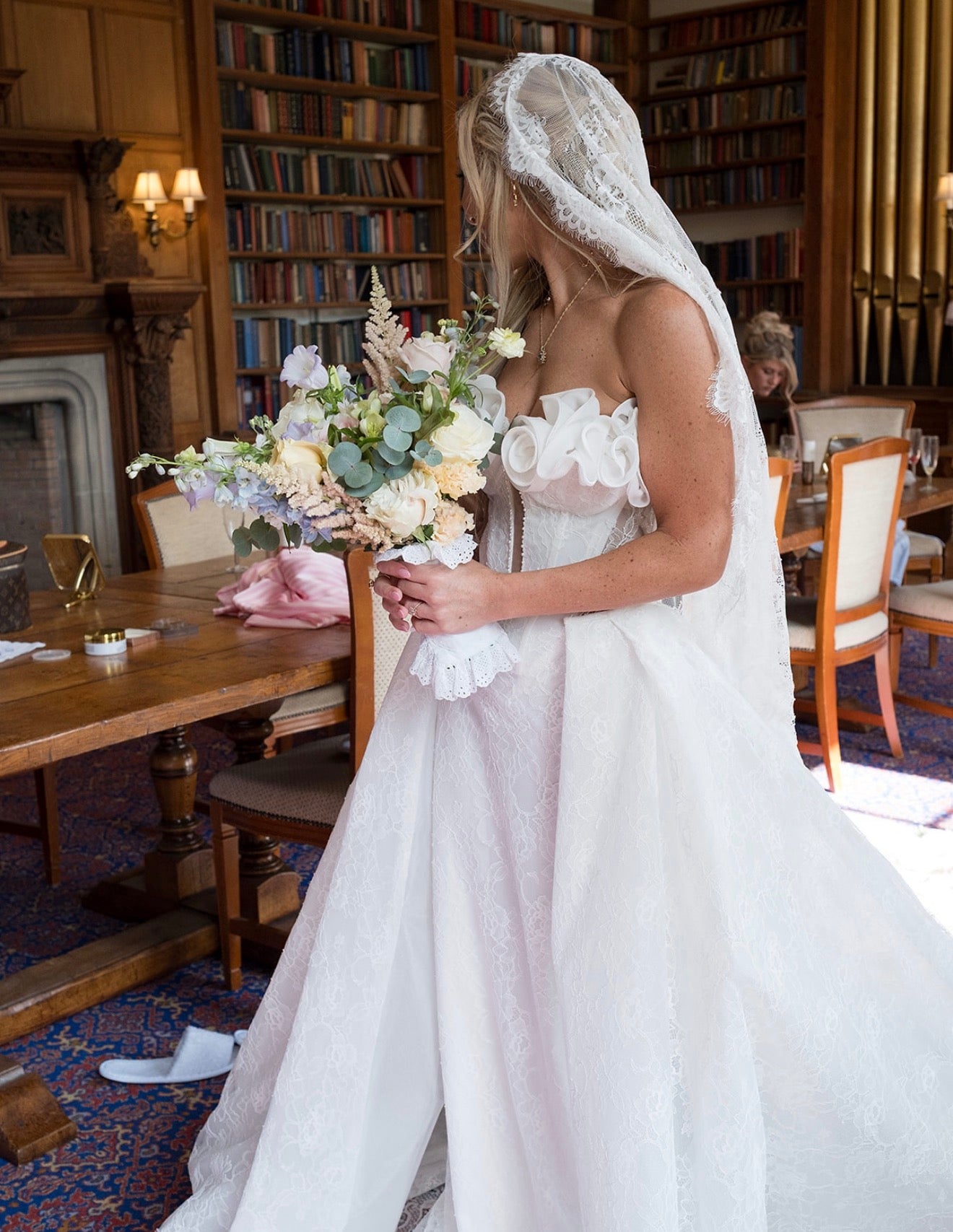 A woman in a strapless white wedding gown with a lace veil holds a large pastel bouquet in a wood-paneled room with bookshelves.