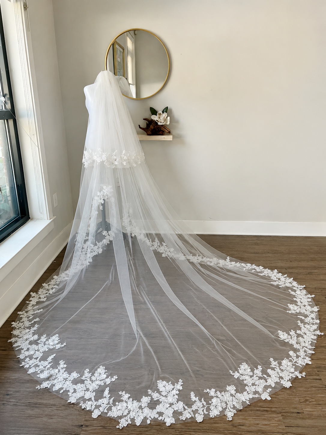 A long white bridal veil with floral lace trim draped over a mannequin in a bright room with wood floors and a round wall mirror.