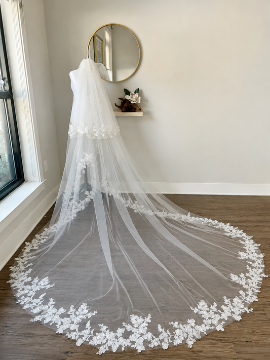 A long white bridal veil with floral lace trim draped over a mannequin in a bright room with wood floors and a round wall mirror.