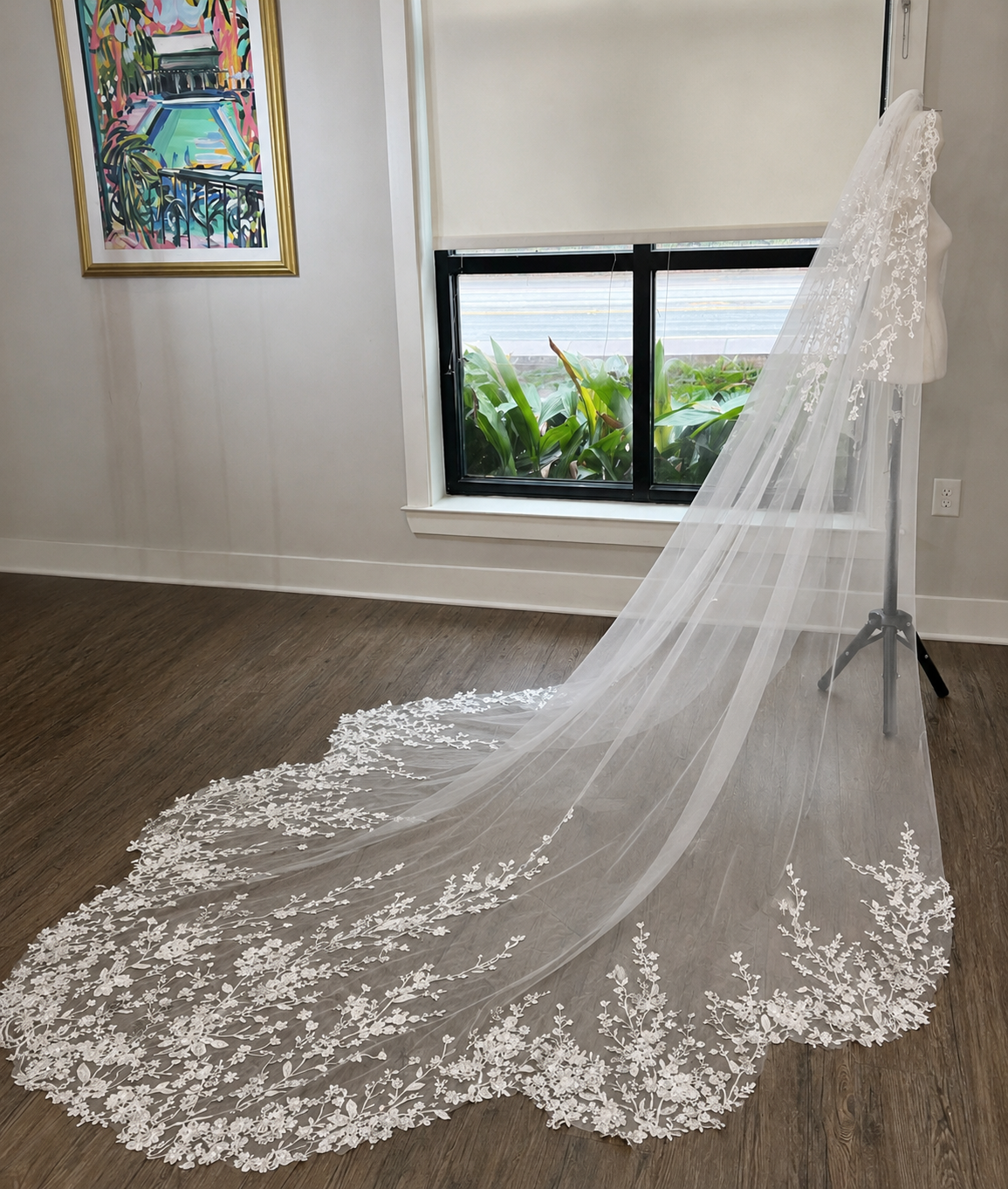 A long white bridal veil with floral lace applique draped across a wood floor in a room, with a mannequin stand visible near a window.