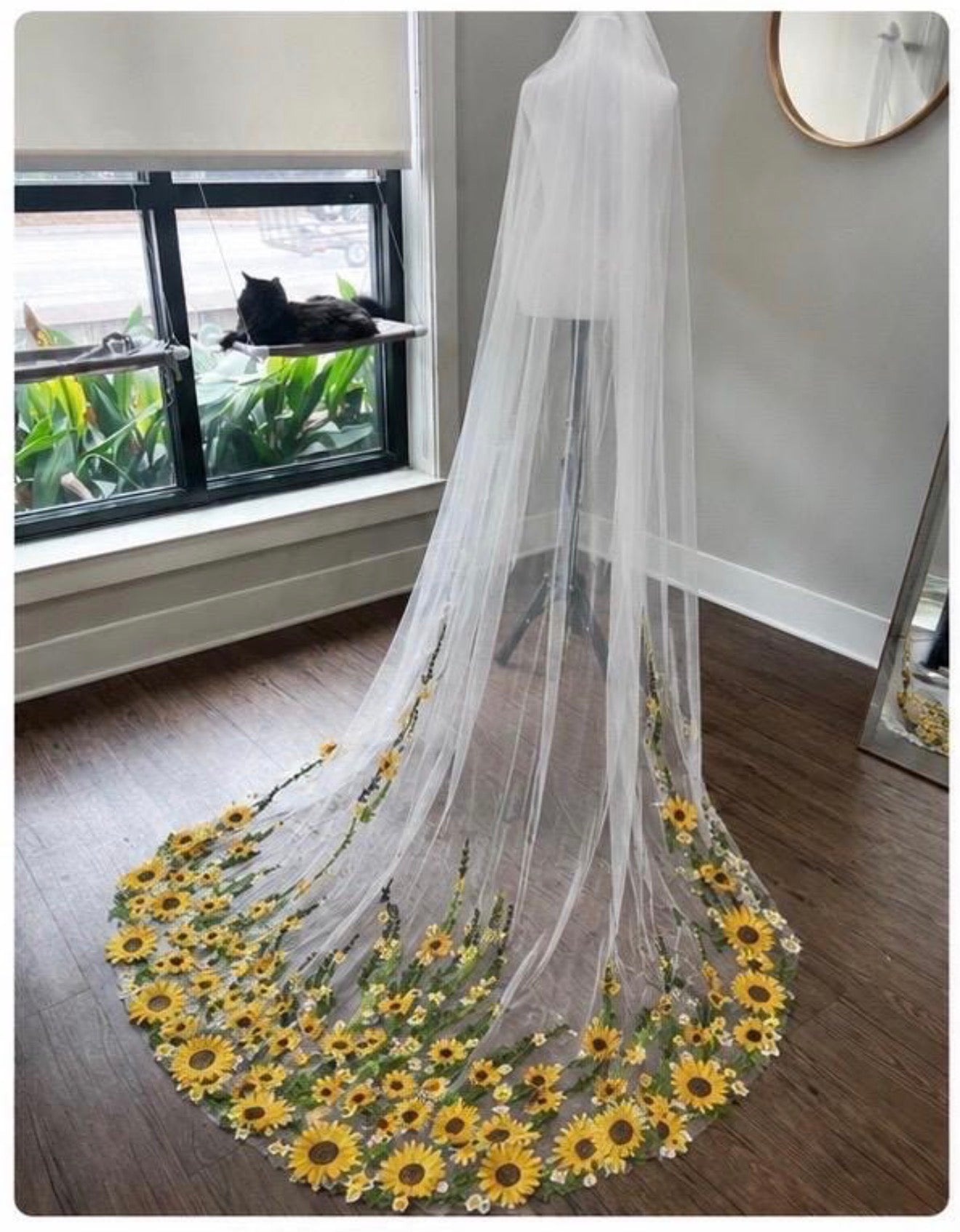 A long white tulle bridal veil with a circular train decorated with embroidered yellow sunflowers and green stems, displayed on a dress form in a room with a window and cat.