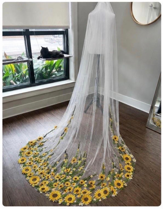 A long white tulle bridal veil with a circular train decorated with embroidered yellow sunflowers and green stems, displayed on a dress form in a room with a window and cat.