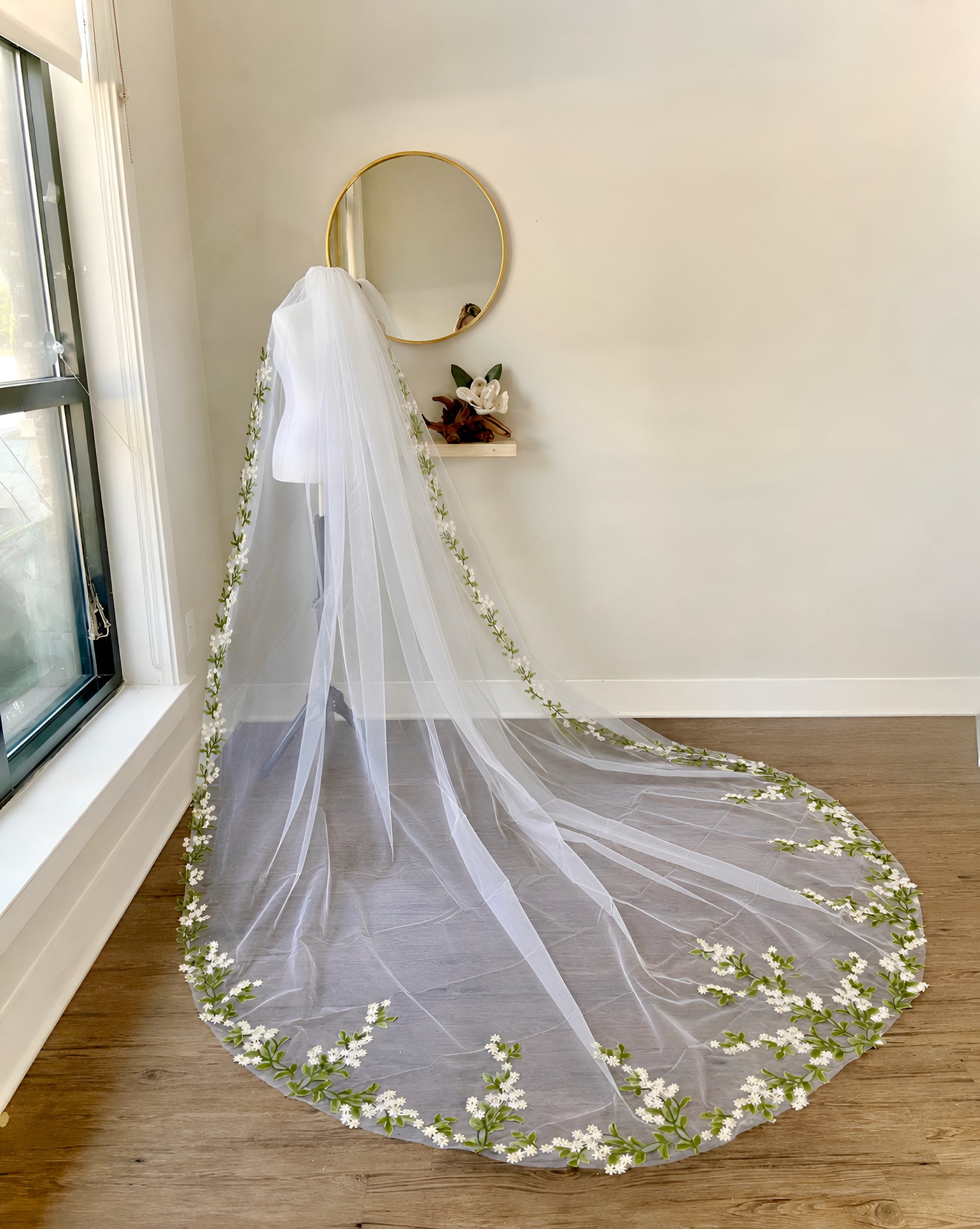 A long white bridal veil with delicate green leafy stems and small white flowers embroidered along the edges, draped over a mannequin in a bright room with a window, wall mirror, and wood floor.