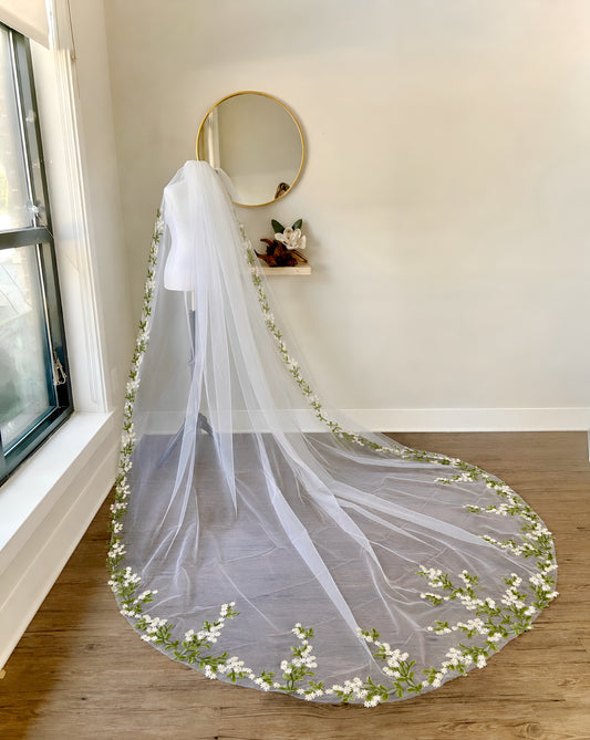 A long white bridal veil with delicate green leafy stems and small white flowers embroidered along the edges, draped over a mannequin in a bright room with a window, wall mirror, and wood floor.